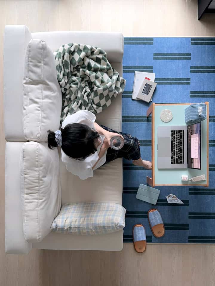 白いソファと青いラグが調和する一人暮らしのリビング、ノートパソコンとブランケットで在宅勤務と読書ができる空間