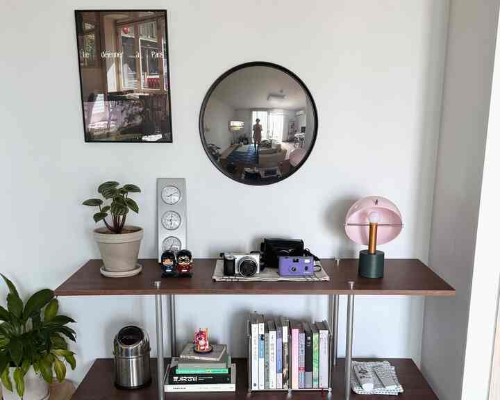 ナチュラルカラーのリビング、木製の棚や植物、丸い鏡とピンクのテーブルランプが配置された雰囲気のあるインテリア