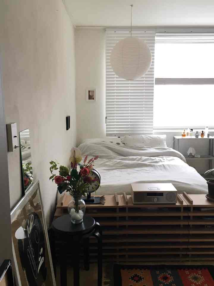 白と木目調が調和した寝室、整ったベッドとブラインド、花瓶が配置された自然な雰囲気のインテリア