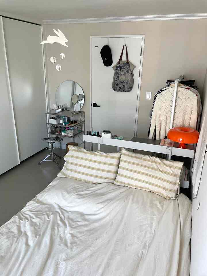 白とグレーのトーンの寝室、シンプルなベッドとストライプの枕、金属製キャビネットに丸い鏡、うさぎのモビールが配置された落ち着いた空間