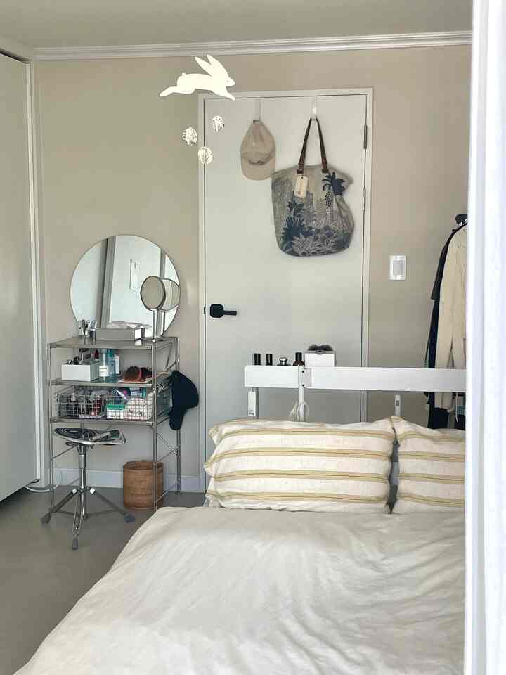 ベージュの壁とホワイトの寝具が調和した落ち着いた寝室、ステンレス製の棚と丸い鏡が配された清潔な空間