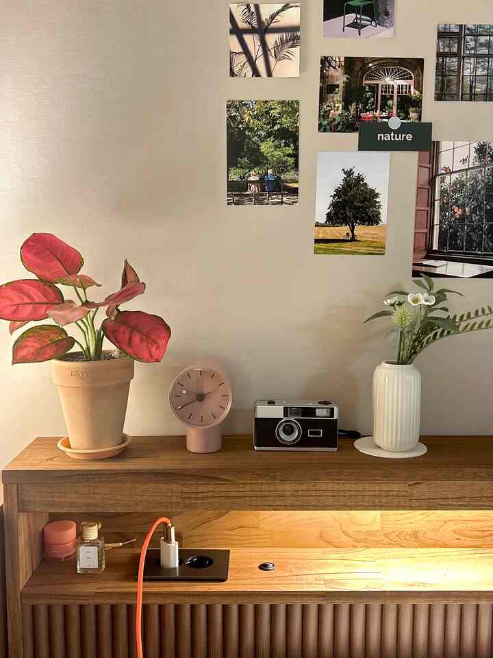 木目調の寝室の壁棚に観葉植物、ピンクの置き時計、フィルムカメラ、造花入りの花瓶、写真のポストカードが調和した雰囲気のある空間