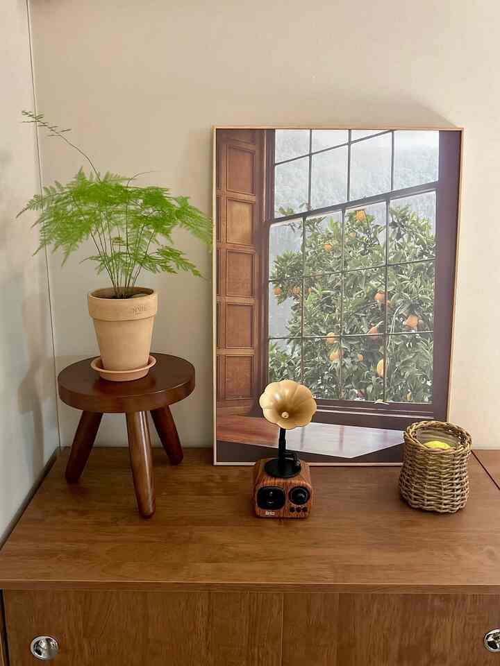 ブラウンの木目調が支配的な空間に、小さな木製スツールと緑の植物、自然なインテリア額縁が配置された雰囲気のある空間