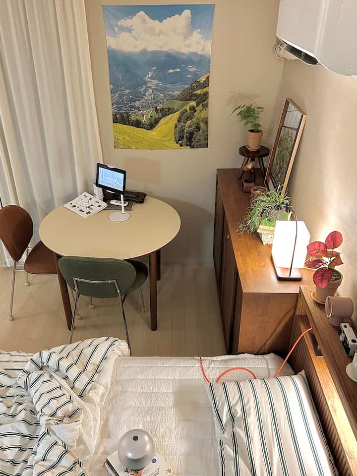 ホワイトとブラウンのトーンのワンルーム寝室、ベッド横のテーブルと棚の植物が配置された落ち着いたナチュラルなインテリア