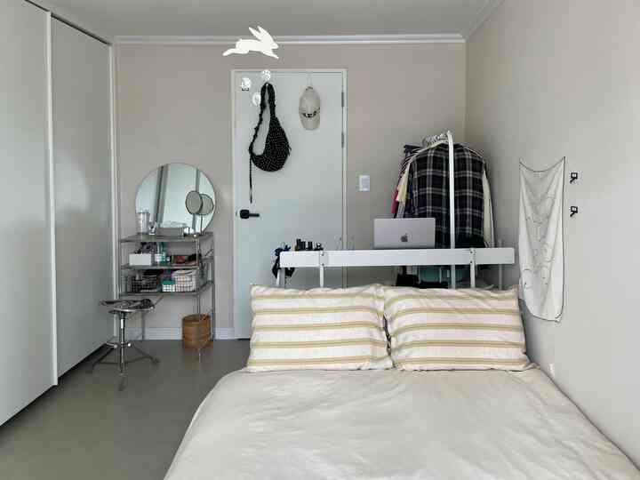 ホワイトとベージュの色調の小さな寝室、ベッドや収納棚、衣類ラックが配置されたミニマルなインテリア