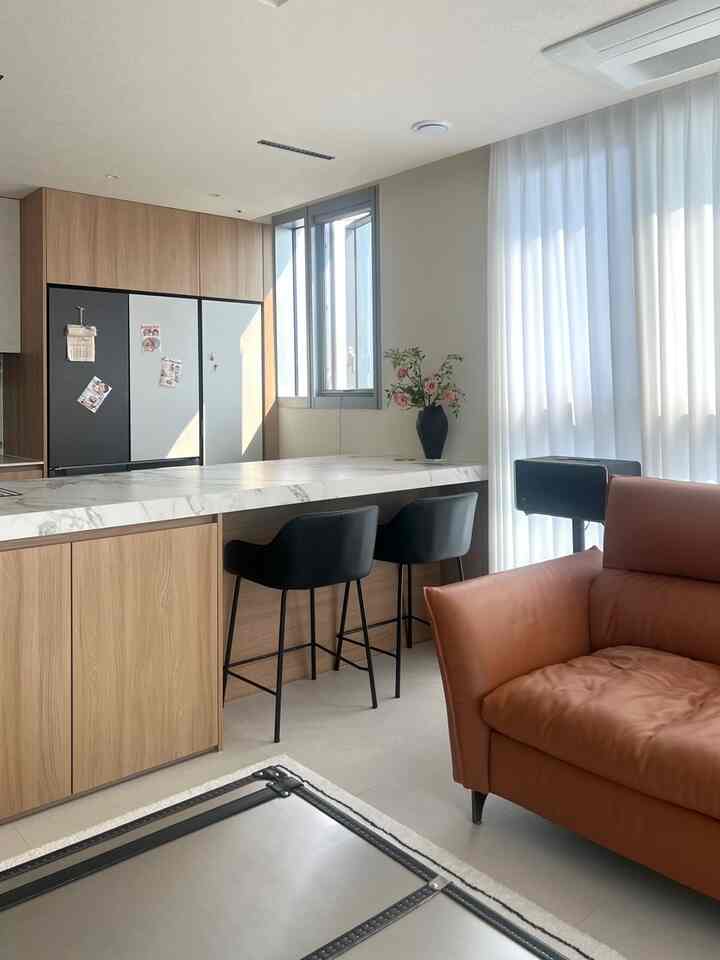 アイボリーと木目調のキッチン兼ホームバー空間、スリムな黒のバースツールとブラウンのレザーソファが配置された雰囲気のあるリビング一体型インテリア