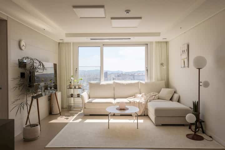 白とベージュのトーンのリビング、コーナーソファと円形のテーブル、フロアランプと植物が配置されたナチュラルモダンなインテリア