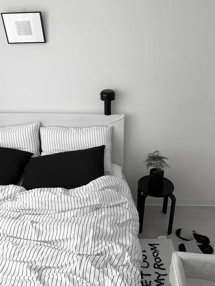 シンプルでモノトーンの寝室、白のストライプ寝具と黒いクッション、黒の丸いスツールとラグが配置された空間
