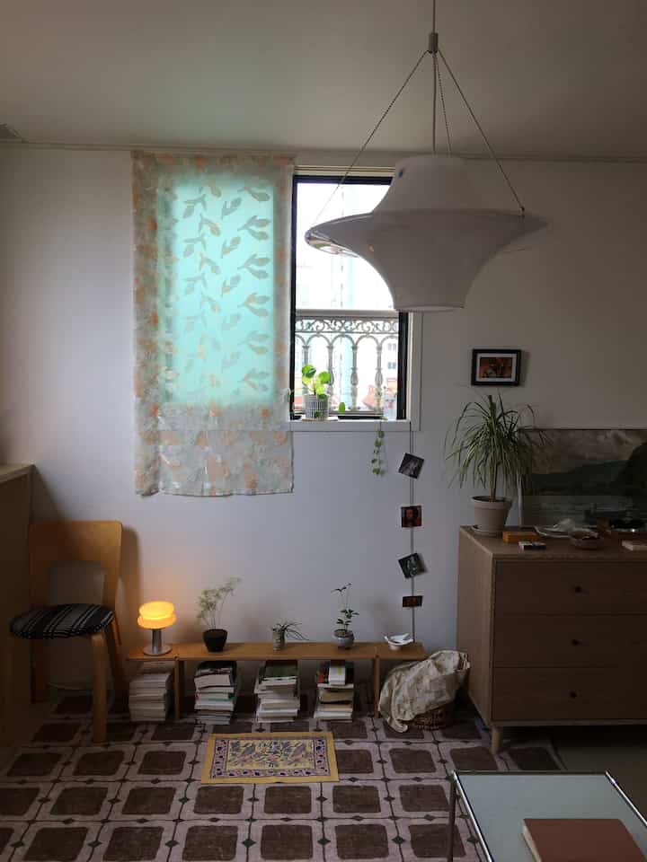 白い壁とナチュラルカラーの家具が調和した書斎、ベンチと本棚、複数の植物、個性的なペンダントライトが配置された雰囲気のあるインテリア