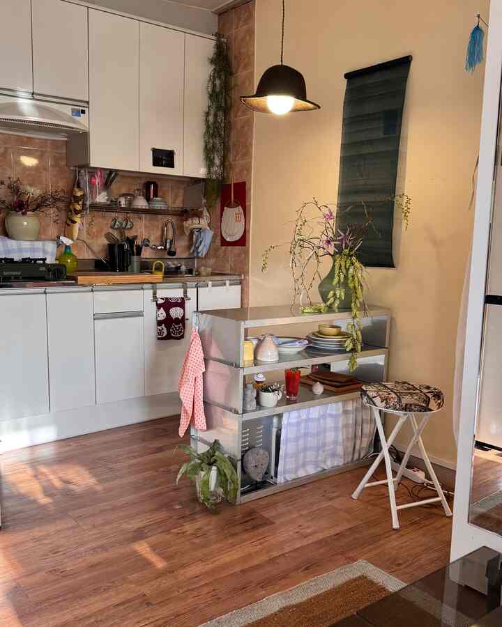 ナチュラルな木目調とホワイトのワンルームキッチン、実用的なメタル収納と折りたたみスツールが配された雰囲気のある空間