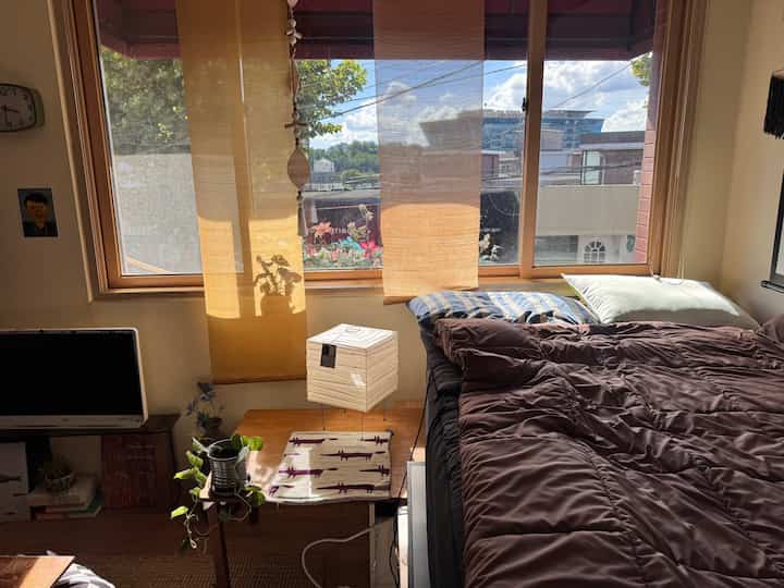 日差しの入るベージュ色のカーテンと濃いブラウンの寝具が配置された落ち着いた寝室