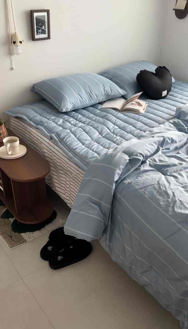 柔らかなブルーの寝具が敷かれた寝室、左側に木製のナイトテーブル、床に置かれた黒いスリッパのある落ち着いた空間