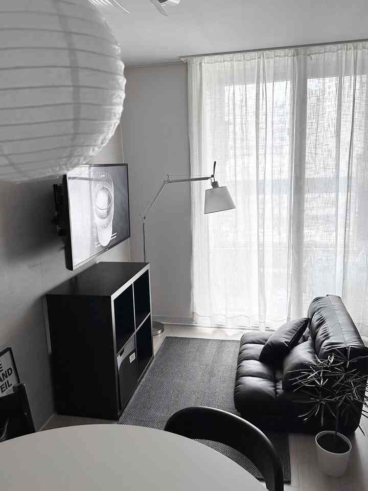 明るい採光と白いカーテンがあるリビングルームに、黒い低めのソファと黒い本棚、フロアランプが配置されたモダンなインテリア