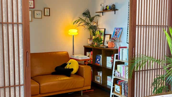温かみのあるナチュラルな色調の小さなリビング、オレンジ色のソファとオーク材の本棚、植物が落ち着いた雰囲気を作り出しているインテリア