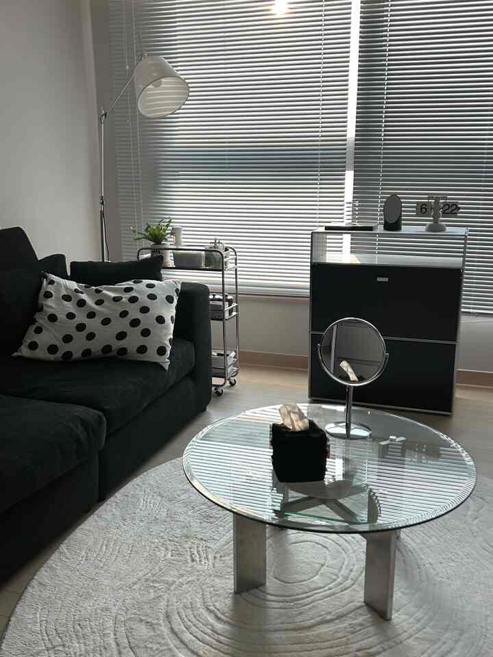 モダンな黒いソファと透明な丸いガラステーブルが配置された明るいリビング、銀色のブラインドとキャスター付きのミッドセンチュリートロリーが見える