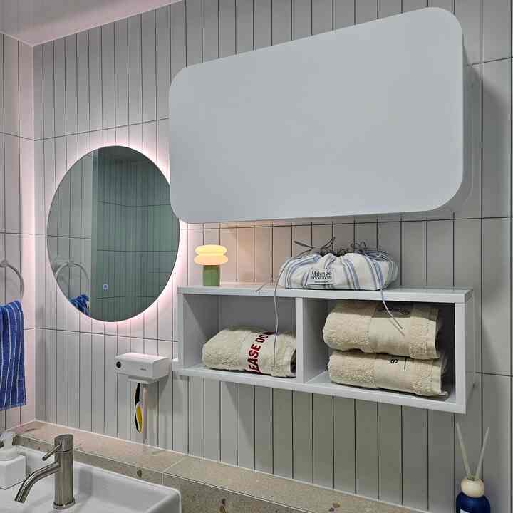 白いタイル壁と木目調の壁掛け収納、LED照明の丸い鏡とタオルが配置された清潔感のある自然なバスルーム