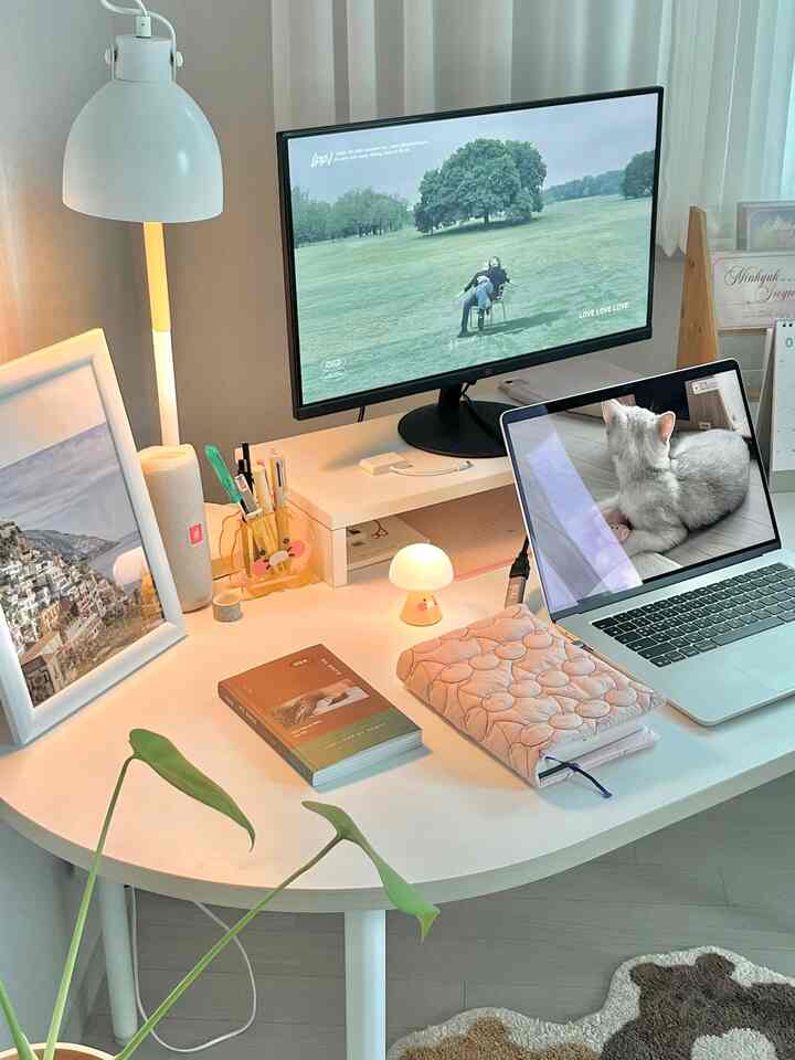 ホワイトトーンのホームオフィス、デュアルモニターとノートパソコン、かわいいペン立てとテーブルランプが置かれた整った作業スペース