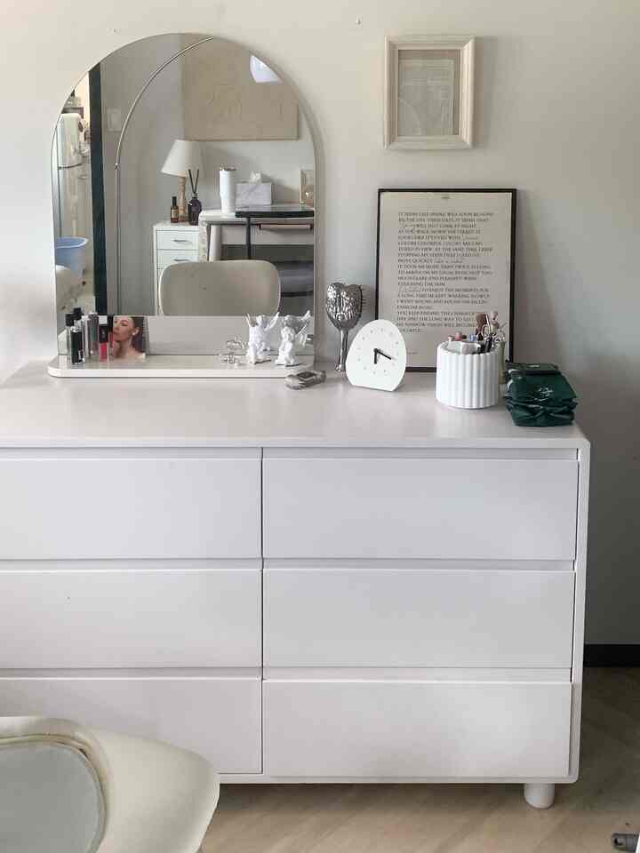 白を基調とした寝室の化粧台空間、ワイドなチェストの上に複数のオブジェ、天使のフィギュア、置き時計、ポスターが配置されたシンプルで清潔感のあるインテリア