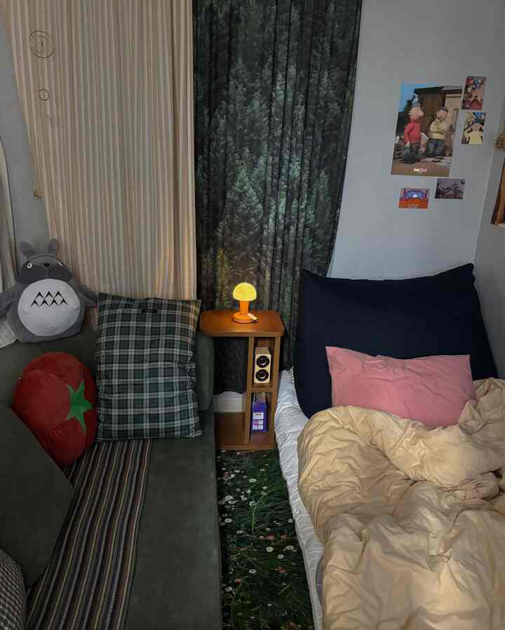 ナチュラルな色調の緑のソファとベッドが配置された、森の柄のカーテンと温かみのある照明が印象的な小さな居心地の良い寝室