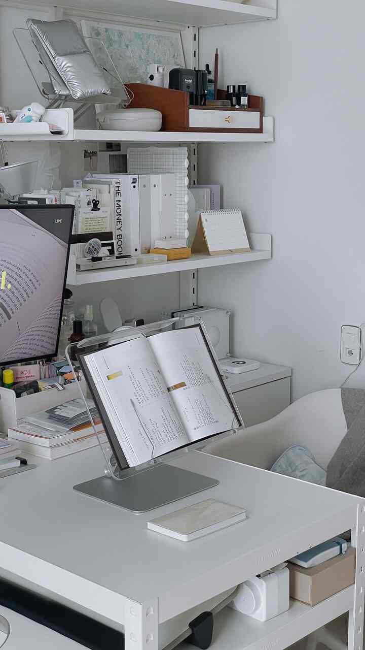 白を基調としたワンルームのホームオフィス、透明なブックスタンドと整頓された収納が配置されたシンプルな作業空間