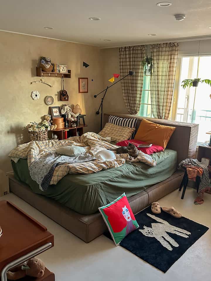 ベージュとグリーンの落ち着いた色調の寝室、中央に大きなベッドと猫、チェックのカーテンとラグが配置された雰囲気のあるインテリア