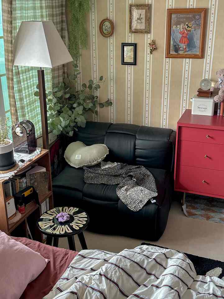 黒いレザーソファと赤いキャビネットがある狭いリビング、ストライプの壁紙と居心地の良い寝具が配置された空間