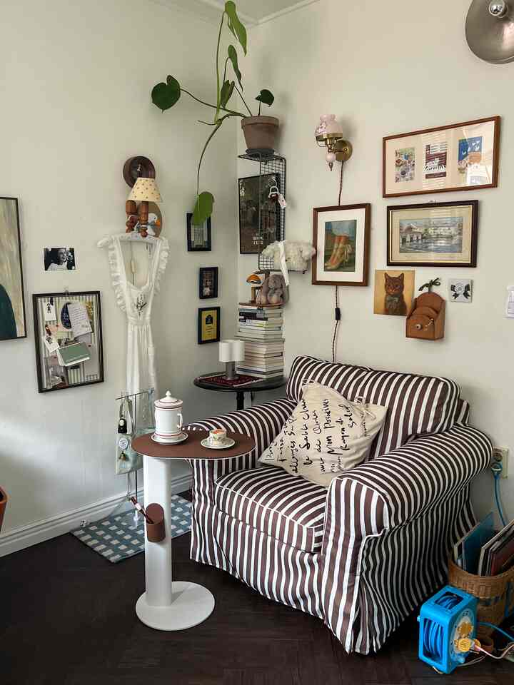 アイボリー色の壁とダークウッドフロアに、ストライプのアームチェアと小さなサイドテーブル、多数のポスターが配置されたナチュラルなヴィンテージ空間