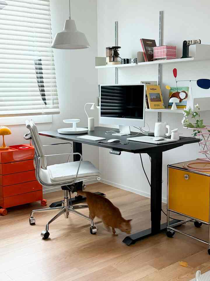 ホワイトとモダン調の書斎、黒いモーションデスクとiMac、オレンジや黄色のアクセント家具が配置された空間