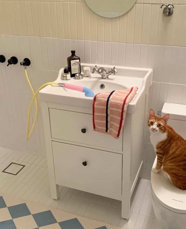 ピンクのタオルと猫がいる白いタイルのバスルーム、洗面台とトイレが配置された雰囲気のある空間