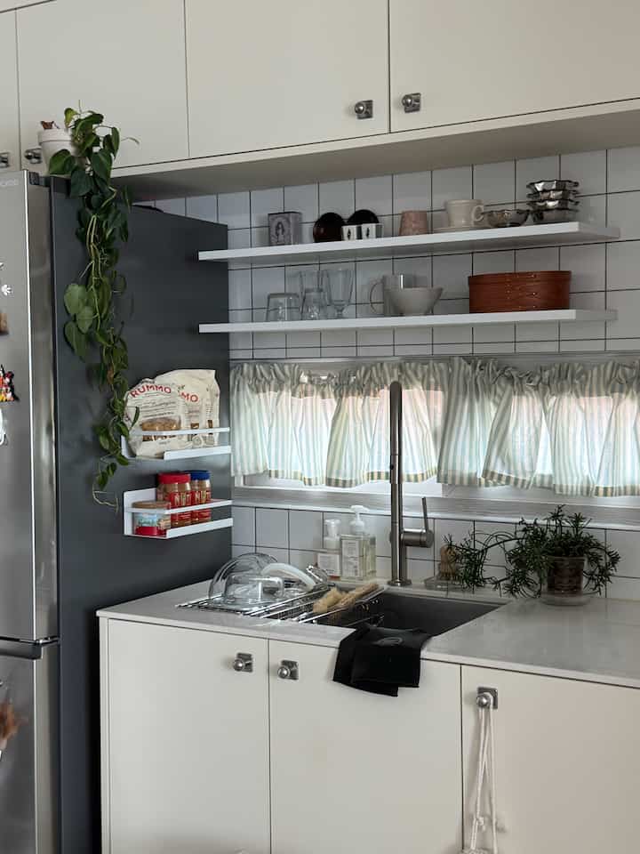 白とグレーのトーンのキッチンで、シンクと食器乾燥ラック、冷蔵庫、ストライプのカーテンが配置された印象的な空間