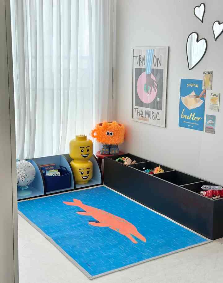 明るい自然光が差し込む白い壁とカーテンの子供部屋、黒い収納箱と青い動物柄のラグ、黄色いレゴヘッドの収納が整った空間