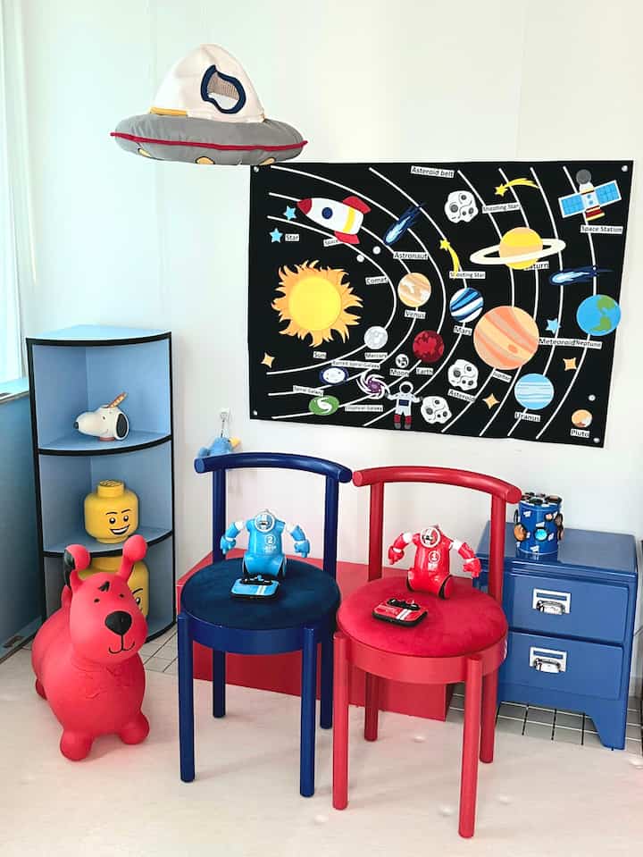 明るい色調の子供部屋、白い壁に赤と青の椅子、宇宙をテーマにした壁飾りと子供のおもちゃが配置された印象的な空間