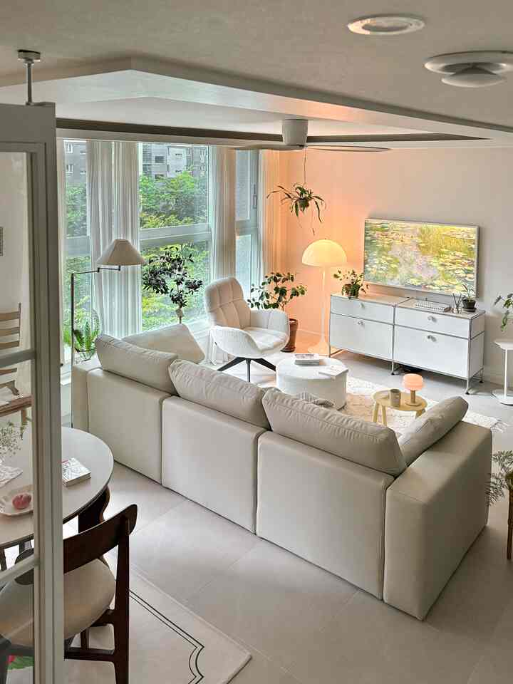 アイボリー調の横長リビング、大きなソファとアームチェア、フロアランプと多様な植物が調和したモダンな空間