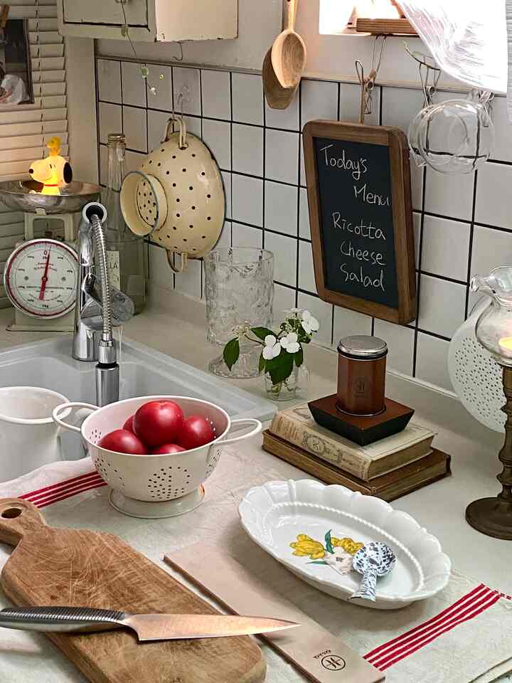 ホワイトのタイル壁と木目調まな板がある明るいキッチンのシンク空間、赤いトマトが入ったホワイトのザルと様々なキッチン用具が配置されているインテリア