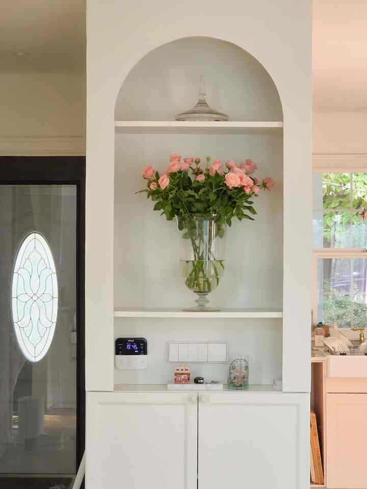 白を基調としたリビングの壁にアーチ状の棚があり、ピンクの花が入った透明なガラスの花瓶が置かれた空間