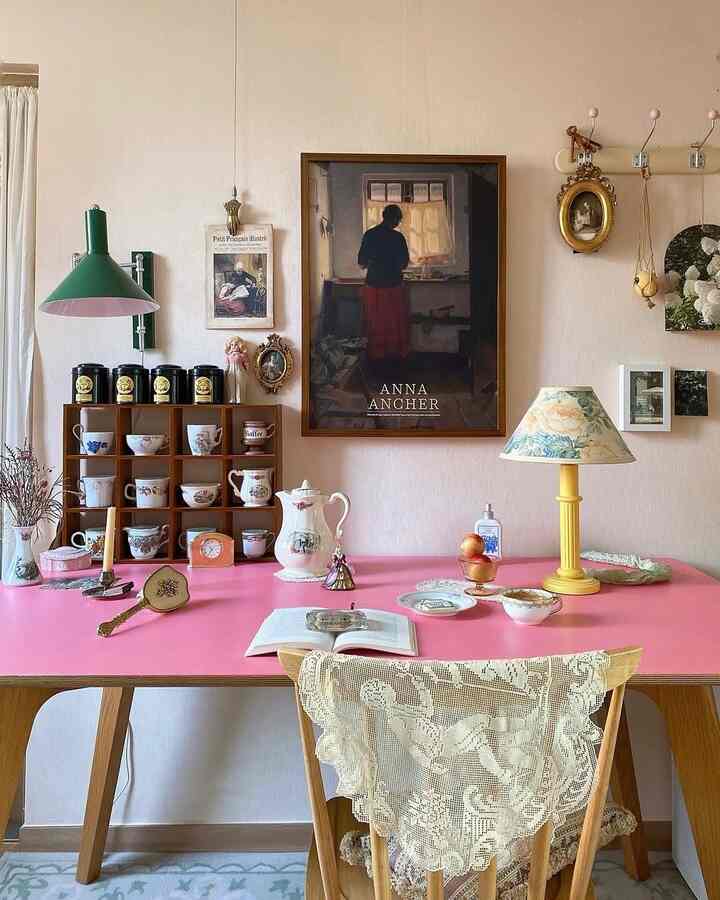 柔らかなベージュの壁とピンクのテーブルがあるヴィンテージなダイニングルーム、絵画とアンティークのキッチン雑貨が配置された落ち着いた空間