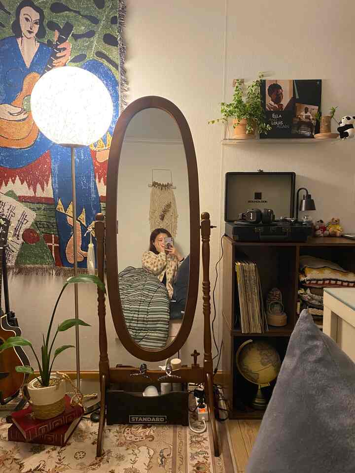 木目調と柔らかい照明が調和する寝室、全身鏡とヴィンテージのレコードプレーヤー、壁デコが配置された雰囲気のあるインテリア