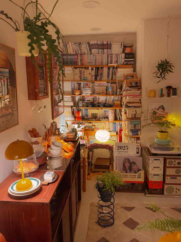暖かい黄色の照明と木製家具、ぎっしり詰まった本棚、多数の植物が配置された居心地の良い小さな読書スペース