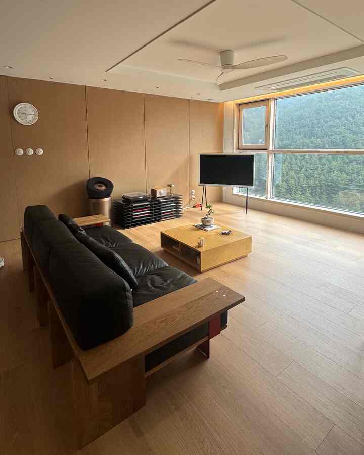 木目調の壁と床、ブラックレザーソファが配置されたモダンでナチュラルなリビング、大きな窓から自然の景色が見える印象的な空間