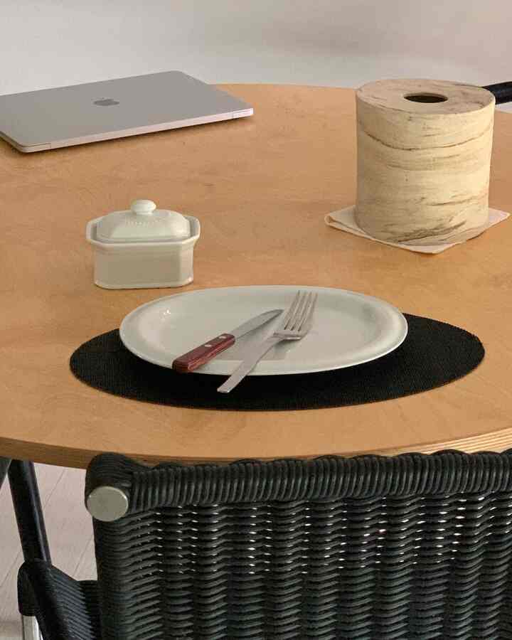 木目調の丸テーブルにノートパソコン、陶器容器、カトラリー、ランチョンマットが配置されたモダンなダイニング空間