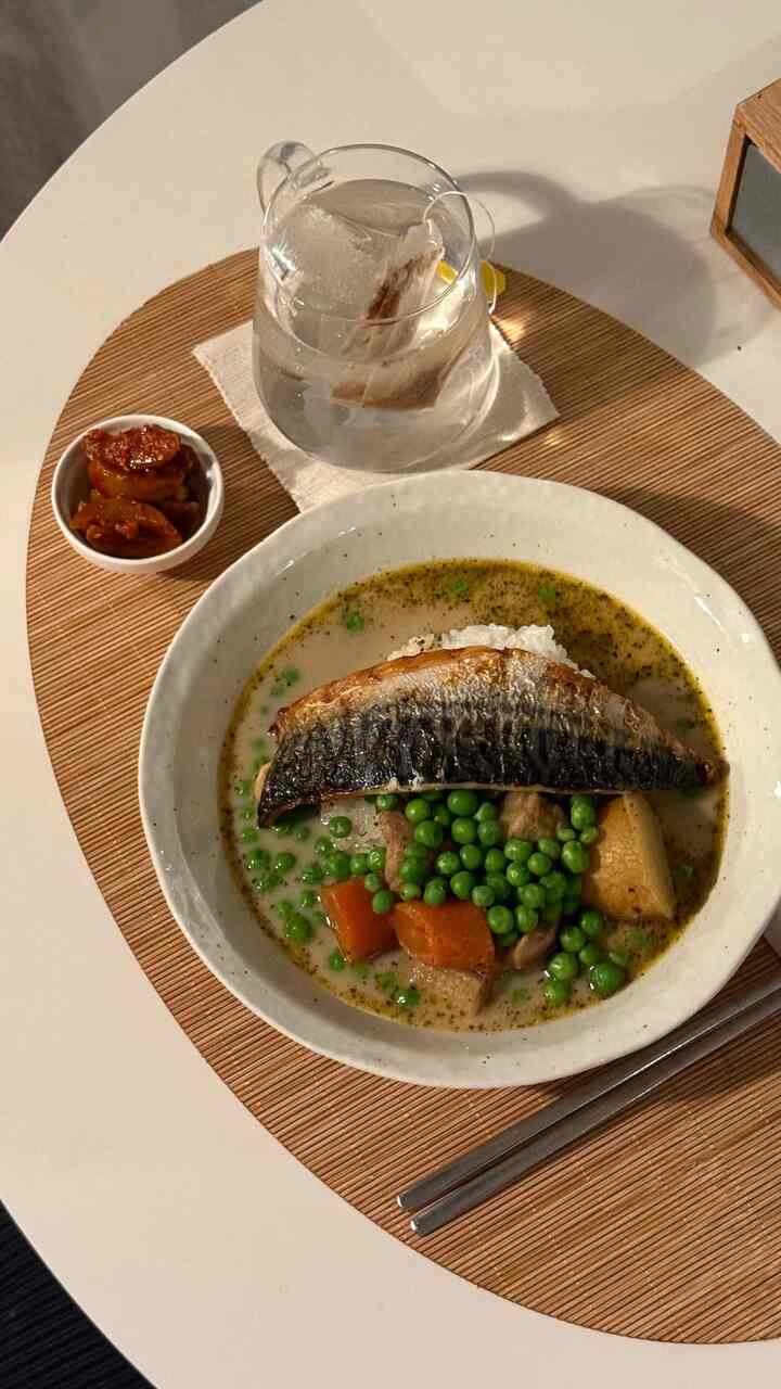 白い丸いダイニングテーブルに木目調のテーブルマットと焼き魚、グリーンピースの食事が配置されたナチュラルモダンな空間