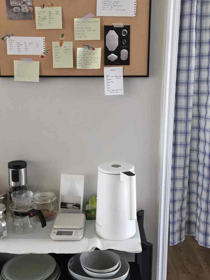 ホワイトの壁とベージュの掲示板があるキッチン、コーヒー用品が置かれたキッチンワゴンと青いチェック柄のカーテンが配置された雰囲気のあるおうちカフェ空間