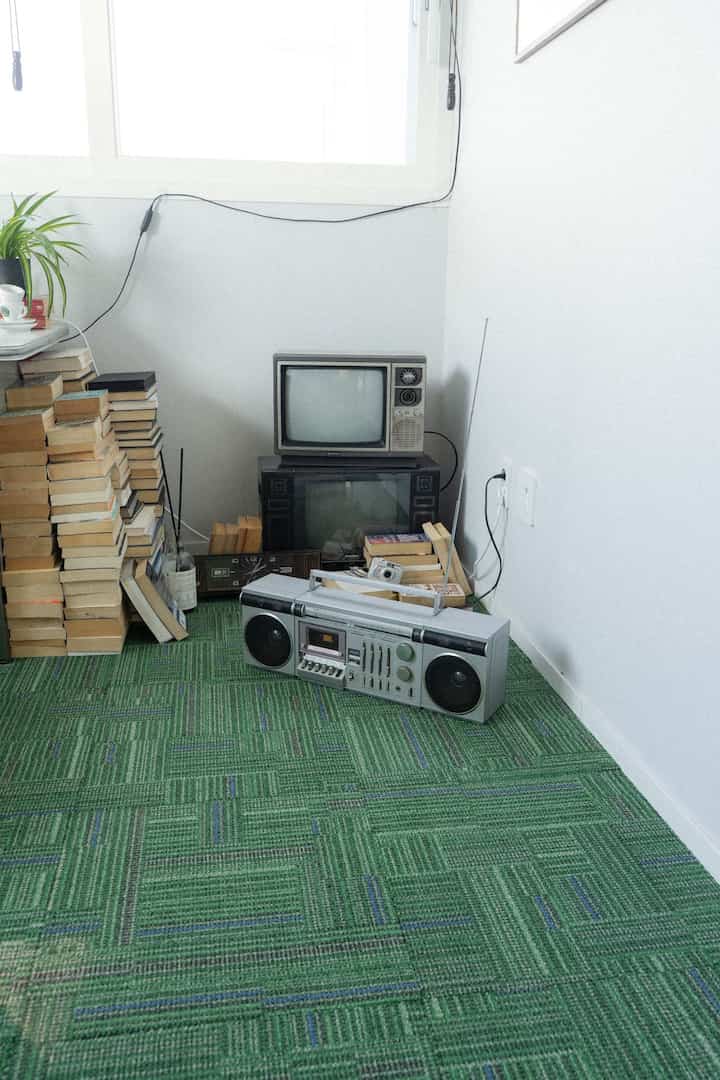 緑のタイルカーペットが敷かれたレトロでヴィンテージ風の空間、古いテレビとラジオ、本の山が置かれた居心地の良いコーナー
