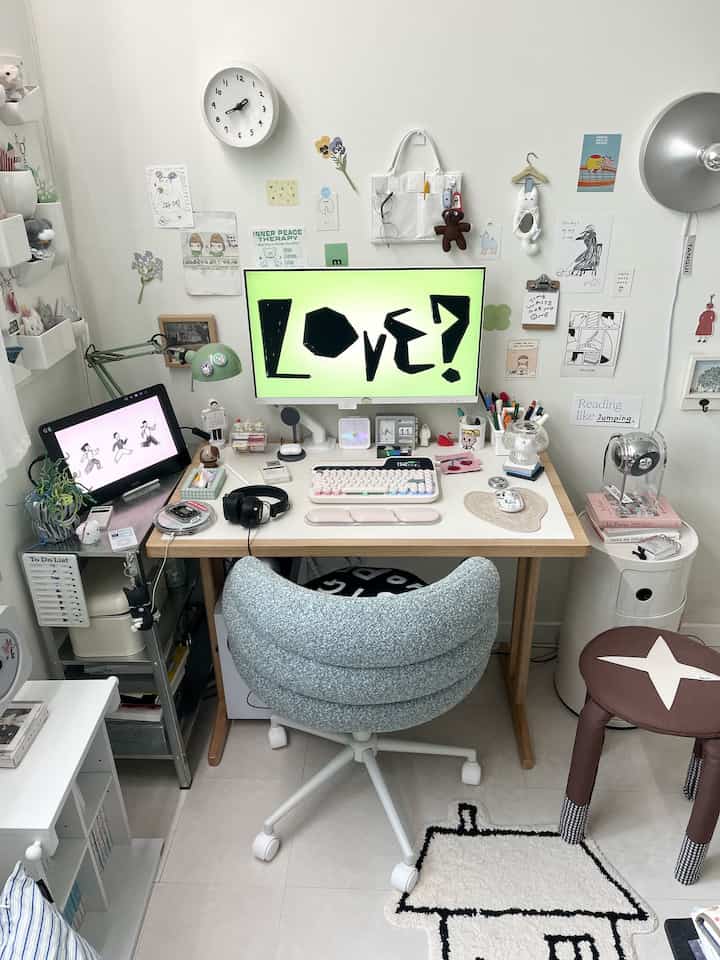 白い壁のコンパクトな部屋に配置された机とブルーグレーの椅子、壁掛け時計と照明がある雰囲気の良いホームオフィス空間