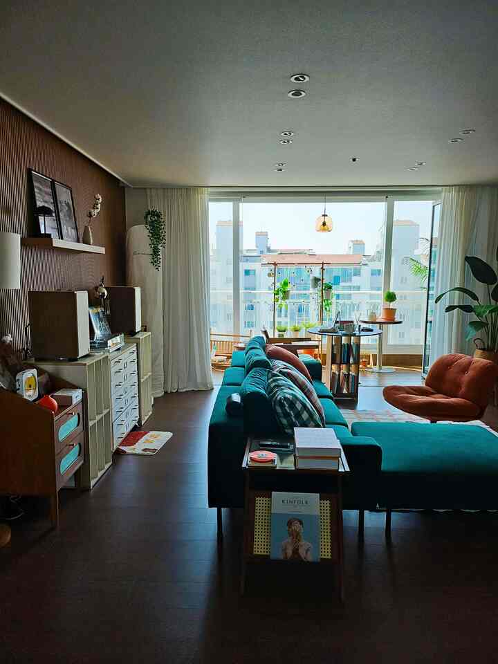 ターコイズグリーンのソファと茶色の木目調の壁が調和したリビング、明るいバルコニーと植物が印象的な空間