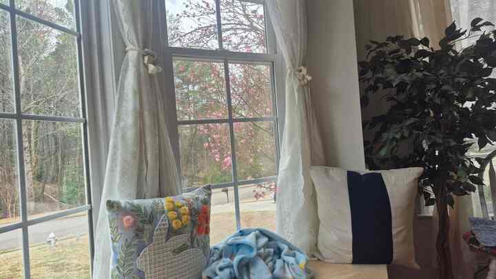 A natural living space with white and beige curtains at the window seat, featuring a blue blanket, floral embroidered cushions, natural light, and indoor plants