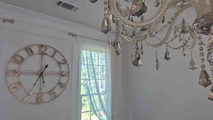 A white and natural-toned dining room featuring a crystal chandelier and large wall clock, creating a traditional and elegant atmosphere