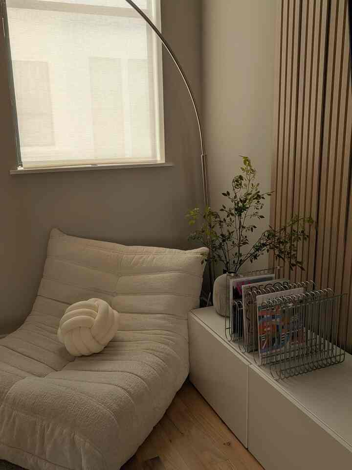 Beige and white toned small cozy corner featuring a soft chair, blinds, floor lamp, and magazine holder with natural interior accents