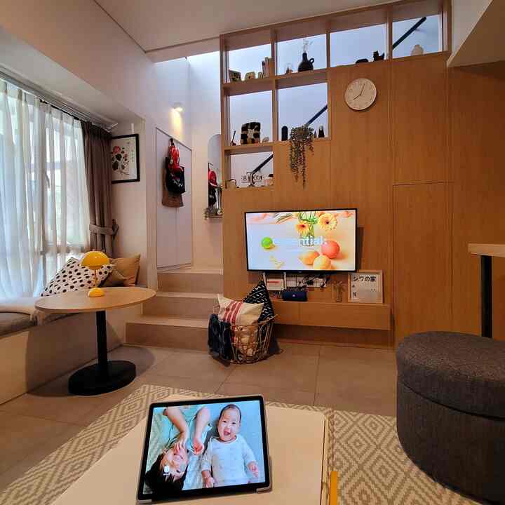 Light brown wood-paneled compact living room with high ceiling, TV, stool, curtains, and children’s photo on tablet creating a cozy atmosphere