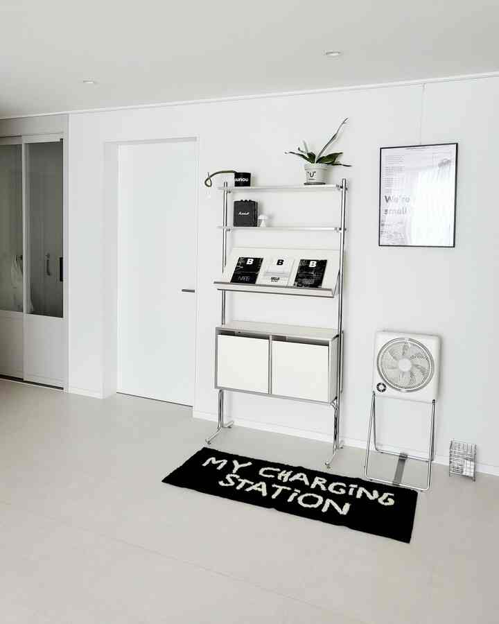 White and silver toned entrance space featuring a simple wall-mounted storage unit and a black rug with white text, creating a clean interior
